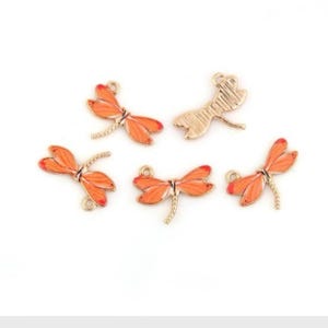 May include: Five gold-colored dragonfly charms with orange enamel. The dragonflies have a detailed design and are perfect for jewelry making or crafting.