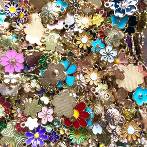 Puede incluir: Un gran surtido de abalorios de flores de metal dorado con detalles de esmalte de colores. Los abalorios presentan una variedad de formas y dise&ntilde;os de flores, incluyendo margaritas, rosas y otros motivos florales.