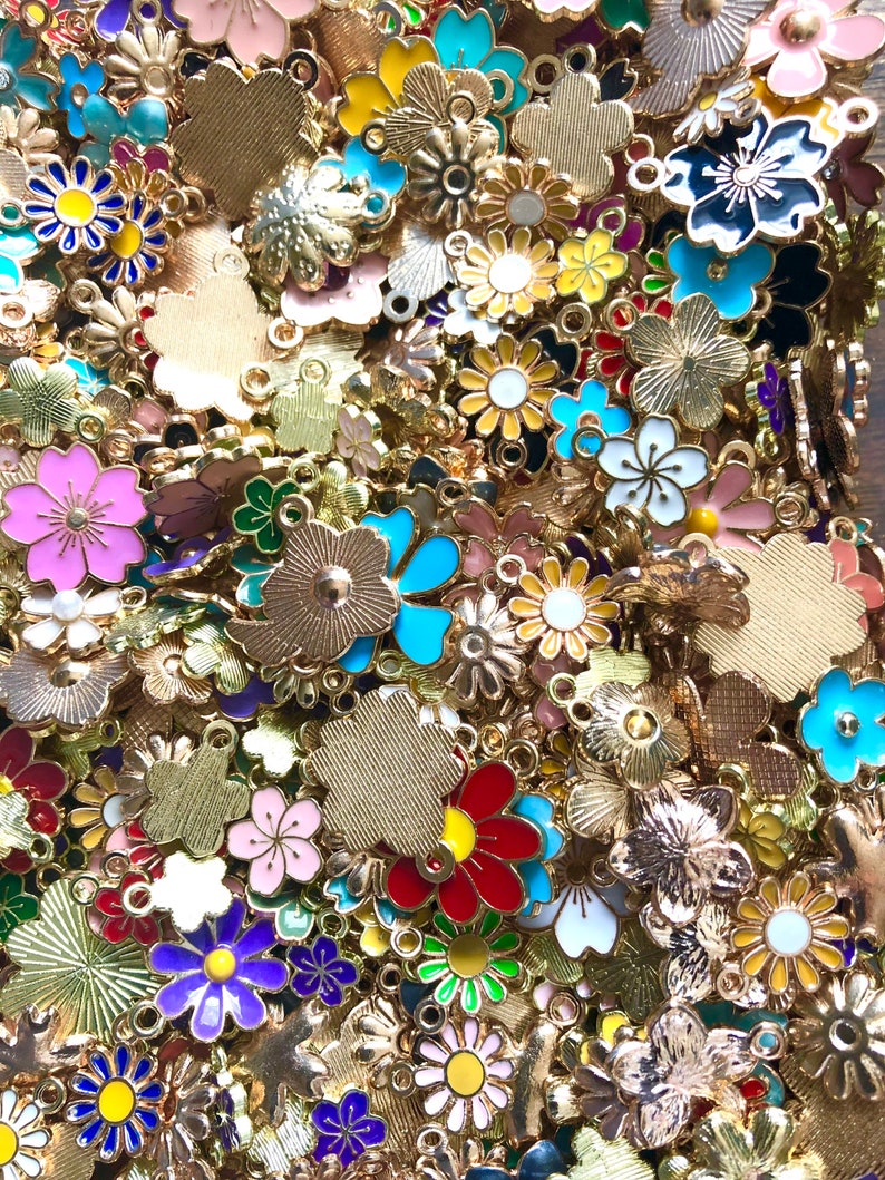 Puede incluir: Un gran surtido de abalorios de flores de metal dorado con detalles de esmalte de colores. Los abalorios presentan una variedad de formas y dise&ntilde;os de flores, incluyendo margaritas, rosas y otros motivos florales.