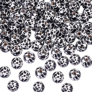 May include: A pile of white beads, each decorated with a black, irregular spotted pattern. The beads are spherical and have a central hole, suitable for stringing. The design is reminiscent of a cow's coat.