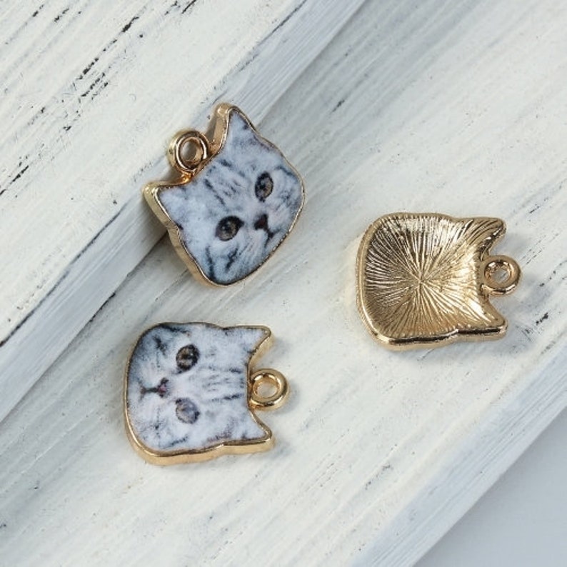 K&ouml;nnte beinhalten: Drei goldfarbene Metall-Anh&auml;nger mit Katzenmotiv. Die Anh&auml;nger haben die Form eines Katzenkopfes und zeigen ein wei&szlig;-graues Katzenbild. Die Anh&auml;nger haben oben eine &Ouml;se zum Anbringen an einer Halskette oder einem Armband.