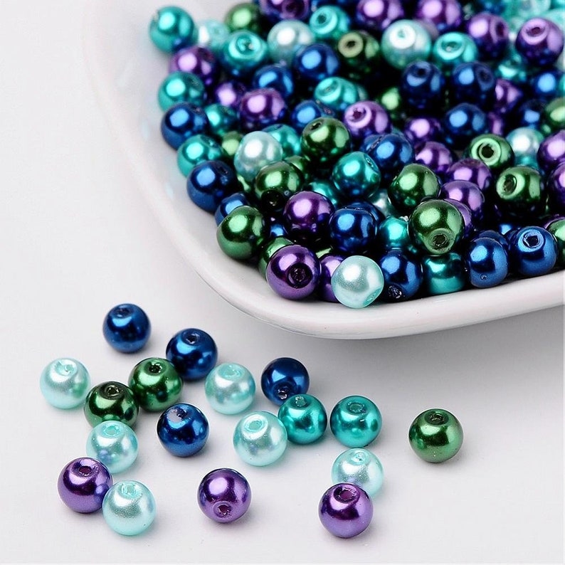 May include: A collection of small, round, iridescent beads in shades of teal, blue, green, and purple. The beads are scattered on a white surface.
