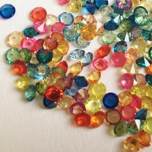 May include: A collection of colorful, faceted glass gems in various shapes and sizes. The gems are scattered on a white surface.