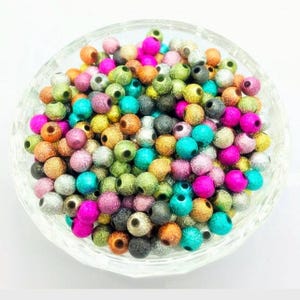 May include: A bowl filled with colorful round beads. The beads are made of wood and have a sparkly finish. The beads are in various colors, including gold, silver, pink, blue, green, and orange.