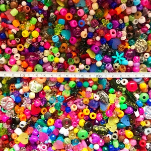May include: A large assortment of colorful plastic beads in various shapes and sizes, including round, square, heart, and star shapes. The beads are arranged in a random pattern and are spread out on a surface. A measuring tape is placed across the top of the image, with the numbers 10, 20, and 30 visible.