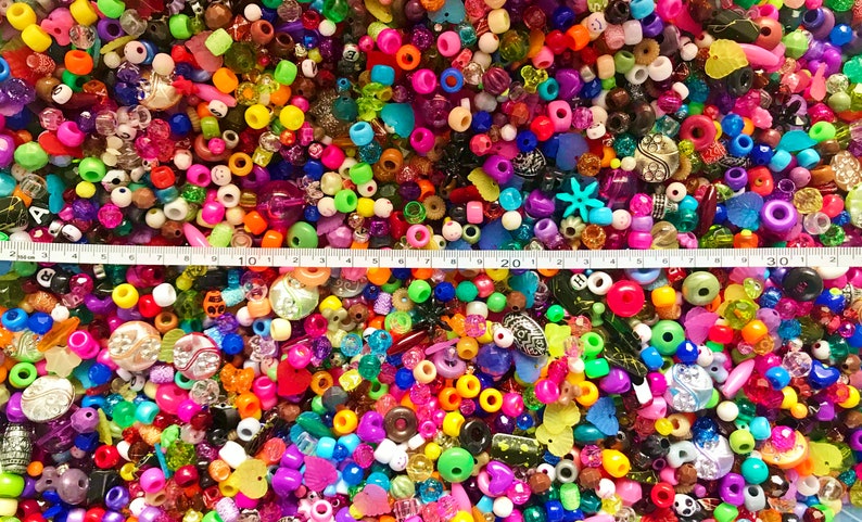 May include: A large assortment of colorful plastic beads in various shapes and sizes, including round, square, heart, and star shapes. The beads are arranged in a random pattern and are spread out on a surface. A measuring tape is placed across the top of the image, with the numbers 10, 20, and 30 visible.