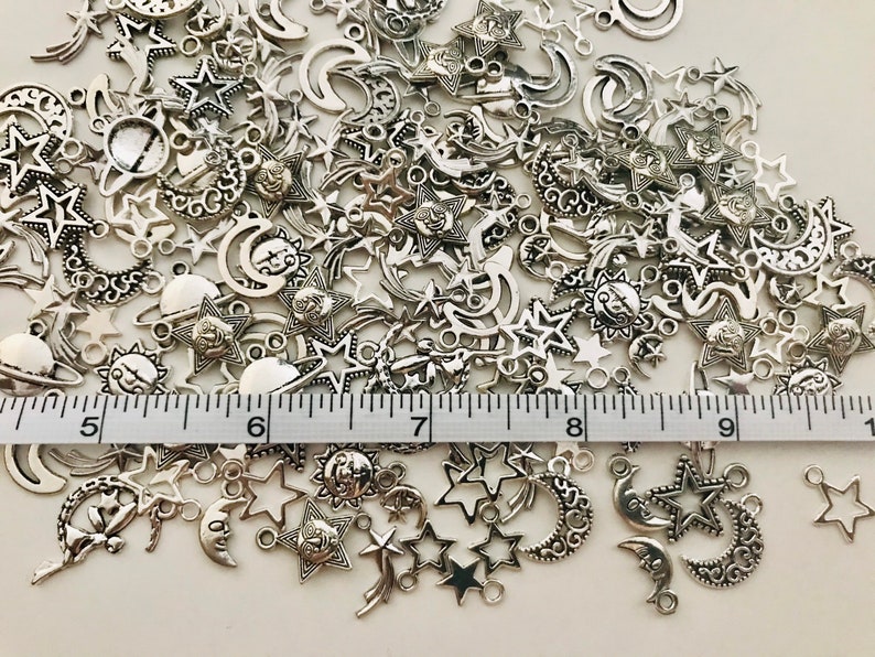 Op de afbeelding: Een grote stapel zilverkleurige metalen bedels in de vorm van sterren, manen en zonnen. De bedels zijn verspreid over een wit oppervlak met een liniaal die 25,4 cm meet.