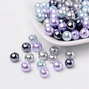 May include: A collection of pearl beads in shades of lavender, light blue, and gray. The beads are arranged in a bowl and scattered on a white surface.
