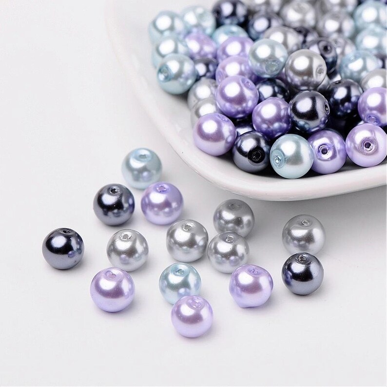 May include: A collection of pearl beads in shades of lavender, light blue, and gray. The beads are arranged in a bowl and scattered on a white surface.