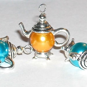 Może przedstawiać: Trzy srebrne breloki w kształcie czajnika z iryzującymi niebieskimi i złotymi wykończeniami. Breloki są szczegółowo wykonane z dzióbkiem, uchwytem i pokrywką.