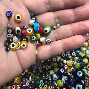 Pu&ograve; includere: Una mano che tiene una variet&agrave; di perline di vetro colorate con un design di occhio turco. Le perline sono di varie dimensioni e colori, tra cui blu, verde, giallo, rosso e bianco.
