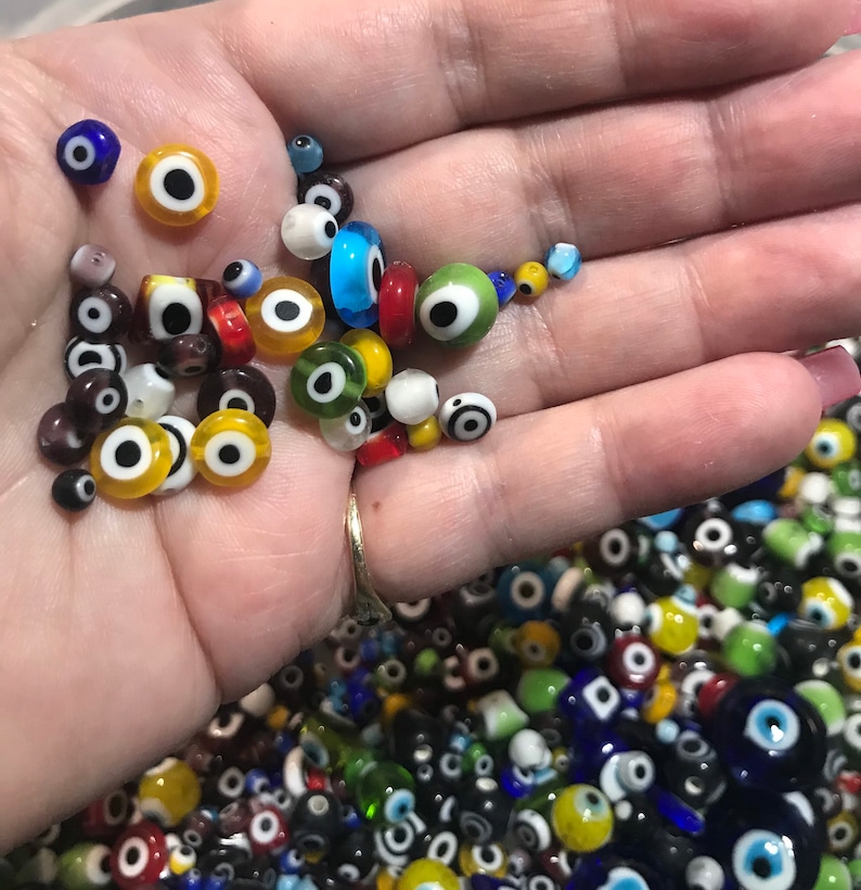 Pu&ograve; includere: Una mano che tiene una variet&agrave; di perline di vetro colorate con un design di occhio turco. Le perline sono di varie dimensioni e colori, tra cui blu, verde, giallo, rosso e bianco.