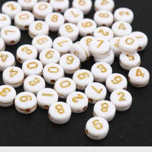 May include: White plastic beads with gold numbers from 0 to 9. The beads are round and have a hole in the center.