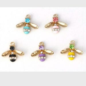May include: Five bee-shaped charms with gold-coloured wings. Each charm has a different coloured body: turquoise, pink, black, purple, and yellow. Each bee has a matching coloured gem on its head and a row of clear gems on its body.