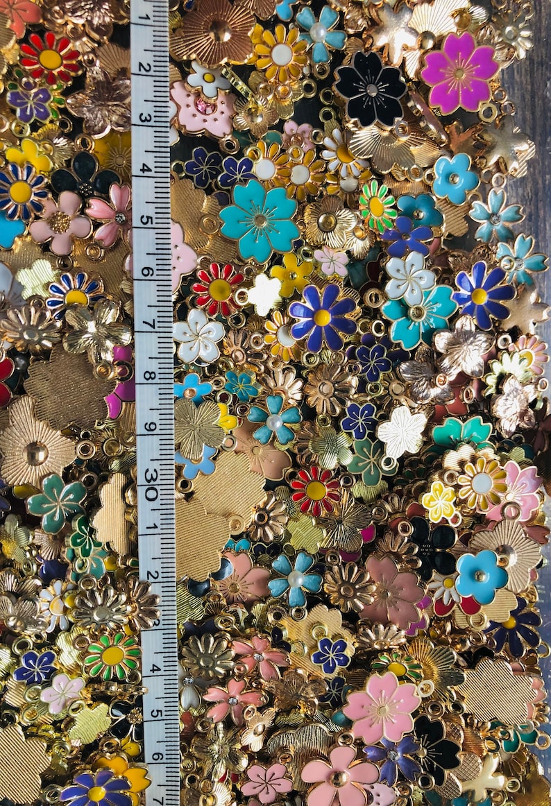 Puede incluir: Un gran surtido de abalorios de flores de esmalte de colores con engastes de metal dorado. Los abalorios est&aacute;n esparcidos sobre una superficie con una regla en el fondo.