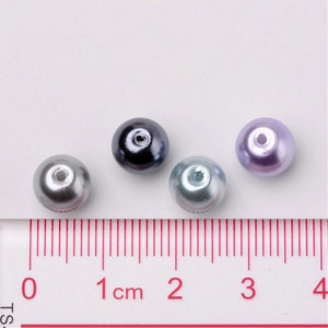 May include: Four round pearl beads in different colors: silver, black, light blue, and lavender. The beads are about 1 cm in diameter and are arranged in a row on a white surface with a ruler for scale.