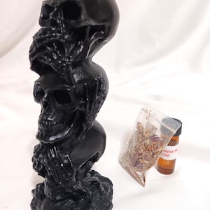 May include: A black candle shaped like three skulls stacked on top of each other. The candle has a textured base and is accompanied by a small bag of herbs and a bottle of oil. The candle is a gothic or Halloween-themed item.