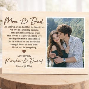 Puede incluir: Un marco de fotos de madera clara con una foto de una pareja. El marco tiene las palabras "Mom & Dad" y un mensaje grabado. Los nombres "Kristen & Daniel" y la fecha "10 de marzo de 2026" también están grabados.