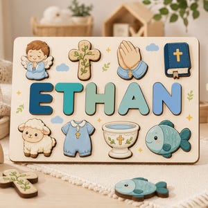 Può includere: Un puzzle in legno con il nome ETHAN in lettere blu e verdi. La tavola include incastri per un angelo in preghiera, una croce, mani giunte, una Bibbia, un agnello, una tutina blu, un fonte battesimale e un pesce.
