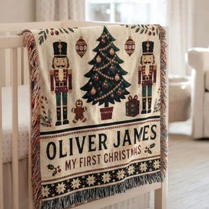 May include: A woven throw blanket featuring a Christmas tree, two nutcrackers, a gingerbread man, and ornaments. The text reads OLIVER JAMES MY FIRST CHRISTMAS. The blanket has a fringed edge and a cream background with red, green, and black details.