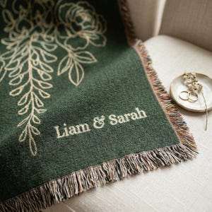 Op de afbeelding: Een donkergroene plaid met een bloemenpatroon en de namen "Liam & Sarah" in het wit geborduurd. De plaid heeft een gefranjerde rand en is over een stoel gedrapeerd.