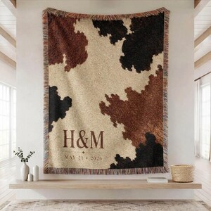 May include: A large, rectangular throw blanket with a cowhide pattern in shades of brown, black, and beige. The blanket has a fringed edge and features the initials "H&M" and the date "MAY 21 2026" printed in a dark brown color.
