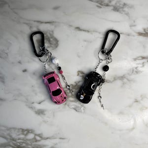 May include: Two car keyrings. One is pink, the other is black. Both have a black carabiner clip, silver rings, and beaded accents in white, pink, and black. Each keyring features a miniature car.