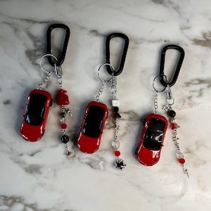 Könnte beinhalten: Drei rote Spielzeugauto-Schlüsselanhänger mit schwarzen Karabinerhaken. Jeder Schlüsselanhänger hat ein rotes Miniaturauto mit schwarzem Dach und dekorative Perlen in Rot, Schwarz und Transparent. Zu den Anhängern gehören Sterne, Blumen und abstrakte Formen.