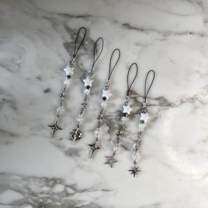 May include: Five phone charms featuring black cord loops, white star beads, clear faceted beads, and silver-toned metal star charms. Each charm has a unique arrangement of small star-shaped links and dangling star pendants in a metallic finish.