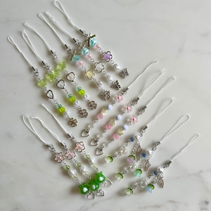 May include: A collection of phone charms featuring silver-toned chains, white cord loops, and various acrylic beads including flowers, hearts, butterflies, and green frogs. Each charm is decorated with small metallic charms and colorful beads.