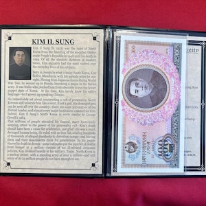 May include: A black binder displaying a portrait of Kim Il Sung and a North Korean 100 Won banknote. The banknote features a portrait and floral design. Text in English and Korean is visible.