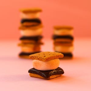 May include: A close-up of a miniature s'more on a pink surface. The s'more consists of a square graham cracker, a toasted marshmallow, and a square of chocolate. Other s'mores are stacked in the background.