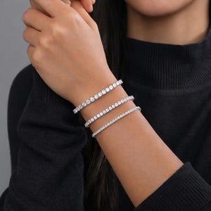 May include: Three diamond tennis bracelets are displayed on a wrist. The bracelets are made of silver metal and feature a row of round, clear diamonds. The person is wearing a black turtleneck sweater.