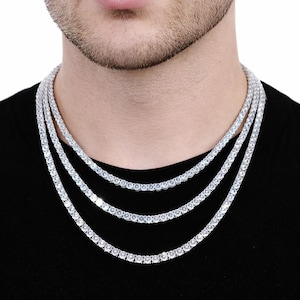 May include: Three diamond tennis necklaces of varying lengths. The necklaces are made of small, round, clear stones set closely together. The necklaces are layered and worn against a black shirt.