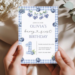 Puede incluir: Una tarjeta de invitación rectangular con un borde de cuadros azules y blancos. La tarjeta presenta arándanos de acuarela y el texto "JOIN US FOR OLIVIA'S berry & first BIRTHDAY". El número "1" también aparece en la tarjeta. El texto adicional incluye fecha, hora, dirección e información RSVP.