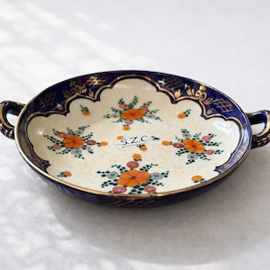 Può includere: Un piatto ovale in ceramica con interno color crema, decorato con motivi floreali arancioni e blu. Il bordo è blu navy con dettagli dorati e ha due piccole maniglie. Le iniziali "S.Z.C." sono visibili.