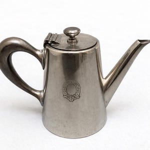 Puede incluir: Una tetera de metal plateado con un asa curva y un pico. La tetera tiene una tapa con bisagras con un pequeño pomo y un emblema decorativo en la parte delantera. La tetera es probablemente vintage o antigua.
