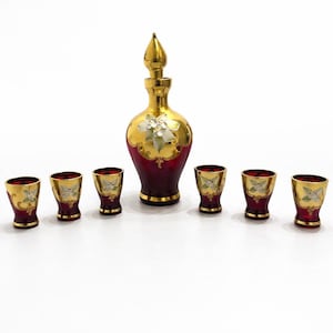 Può includere: Un set di decanter rosso e oro vintage con sei bicchierini abbinati. Il decanter ha un tappo dorato e un motivo floreale. I bicchierini e il decanter hanno un bordo dorato.