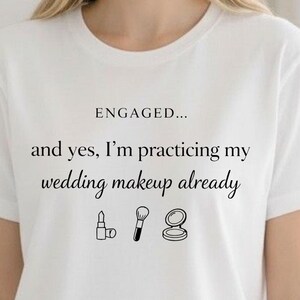 May include: A white short-sleeved t-shirt featuring the black text ENGAGED... and yes, I'm practicing my wedding makeup already, accompanied by small black line art icons of a lipstick, a makeup brush, and a compact powder case.