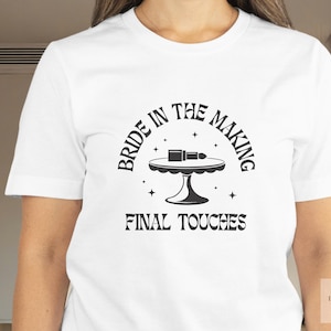 May include: White t-shirt with the text "BRIDE IN THE MAKING FINAL TOUCHES" in black. The design features a table with makeup items. The shirt is a short-sleeved crew neck.