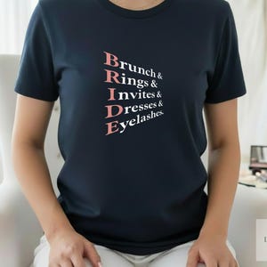 May include: Navy blue t-shirt with the word "BRIDE" in pink, with the words "Brunch & Rings & Invites & Dresses & Eyelashes" printed above and below. The shirt is a crew neck and is being worn.