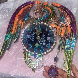 May include: A decorative wall clock with a dragon wing design. The clock face is dark blue with crystal accents and gold hands. The wings are orange, purple, and green with glitter. A matching pendant hangs below.