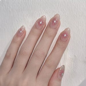 May include: Oval-shaped fingernails featuring a sheer, translucent base coat with a soft pink gradient at the center of each nail. Each nail is decorated with a delicate, silver glitter starburst design that radiates outward from the center point.