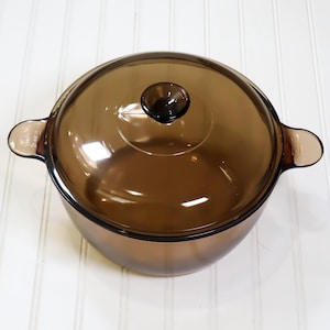 May include: A round, translucent brown glass cooking pot with a matching glass lid. The pot features two integrated side handles and a central knob on the lid. The glass has a smooth, polished finish, allowing light to pass through the dark amber material.