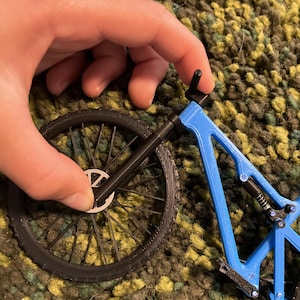 Puede incluir: Un modelo en miniatura de bicicleta azul y negra. El cuadro es de un azul vibrante, contrastando con las ruedas y otras piezas negras. Una mano sostiene el modelo, mostrando su dise&ntilde;o detallado.