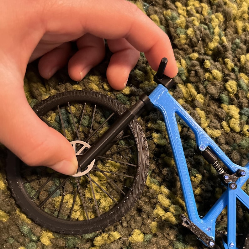 Puede incluir: Un modelo en miniatura de bicicleta azul y negra. El cuadro es de un azul vibrante, contrastando con las ruedas y otras piezas negras. Una mano sostiene el modelo, mostrando su dise&ntilde;o detallado.