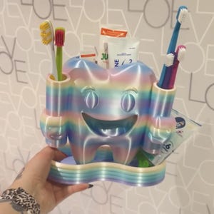 May include: A rainbow-colored tooth-shaped toothbrush holder with a smiling face. The holder has two holes for toothbrushes and a tray for toothpaste and other dental products. Several toothbrushes and toothpaste are visible.