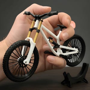 Könnte beinhalten: Ein Miniatur-Mountainbike-Modell mit weißem Rahmen, schwarzen Reifen mit detailliertem Profil, schwarzem Lenker und goldfarbenen Vorderradgabeln. Das kleine Fahrrad verfügt über einen hinteren Stoßdämpfer und einen schwarzen Kunststoffständer.