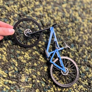 Puede incluir: Un modelo de bicicleta peque&ntilde;o, azul y negro, con dos ruedas y un cuadro detallado. La bicicleta est&aacute; sujeta por una mano, mostrando el intrincado dise&ntilde;o de la bicicleta de monta&ntilde;a en miniatura. Las ruedas tienen neum&aacute;ticos negros y radios plateados.