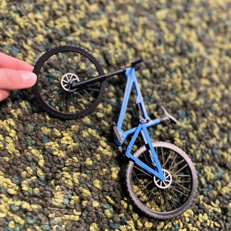 Puede incluir: Un modelo de bicicleta peque&ntilde;o, azul y negro, con dos ruedas y un cuadro detallado. La bicicleta est&aacute; sujeta por una mano, mostrando el intrincado dise&ntilde;o de la bicicleta de monta&ntilde;a en miniatura. Las ruedas tienen neum&aacute;ticos negros y radios plateados.
